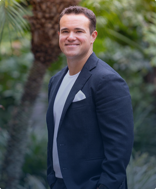 Smiling man wearing a dark blazer and white t-shirt standing outdoors with green foliage background.