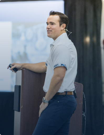 Man wearing a white shirt with rolled-up sleeves and a headset microphone giving a presentation while standing beside a podium.