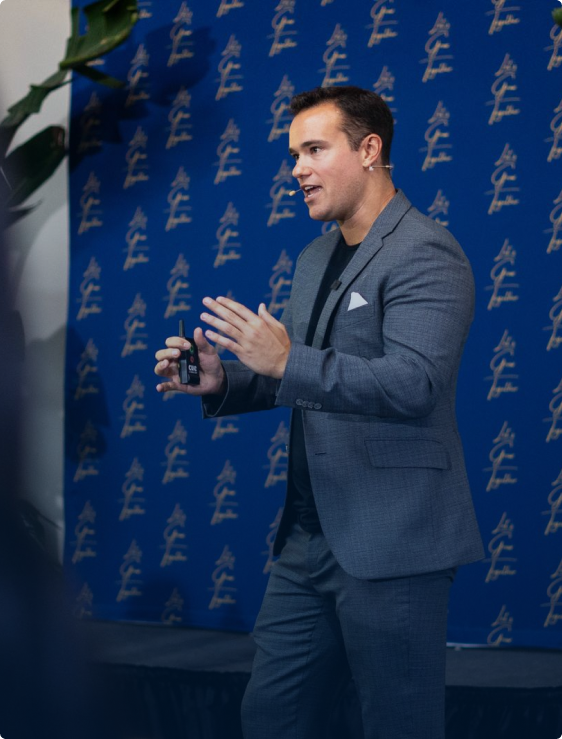 Man in a gray suit speaking on stage with a microphone headset and holding a small bottle in front of a blue backdrop with repeated gold text.