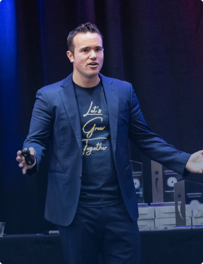 Man in a navy blazer and t-shirt saying 'Let's Grow Together' speaking on stage with awards on a table behind him.