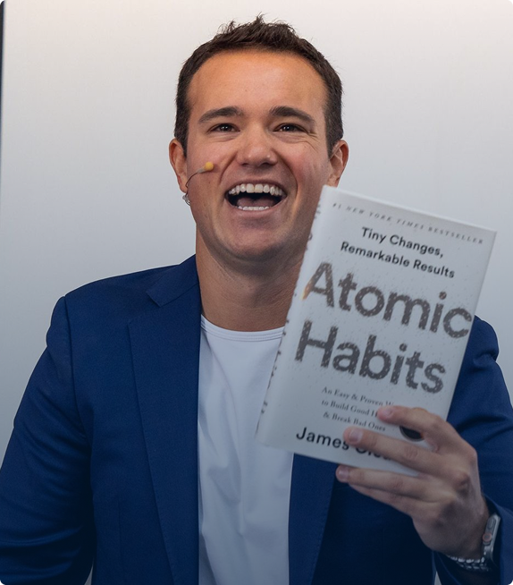 Smiling man in a blue blazer holding the book 'Atomic Habits' by James Clear.