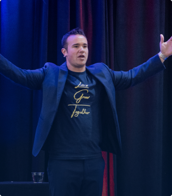 Man wearing a blazer and a navy shirt with 'Live Grow Together' text, speaking with arms outstretched on stage.