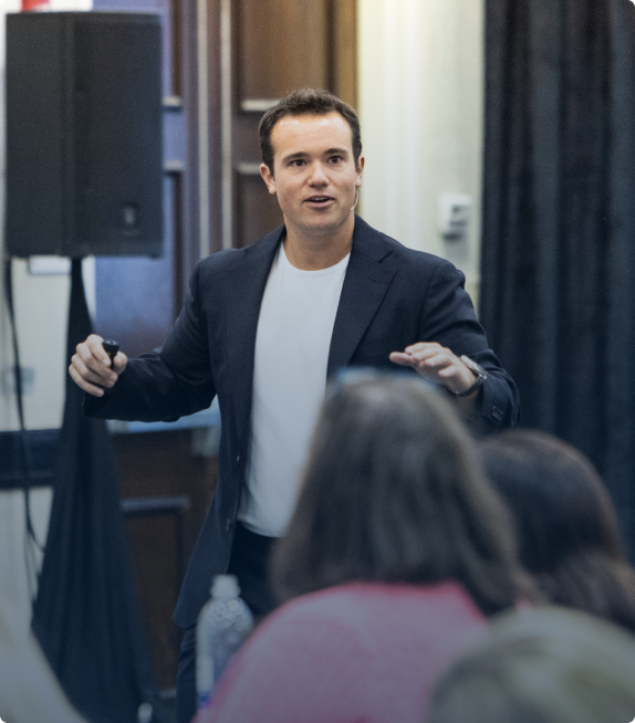 Man in a black blazer and white shirt speaking and gesturing during a presentation with audience members blurred in the foreground.