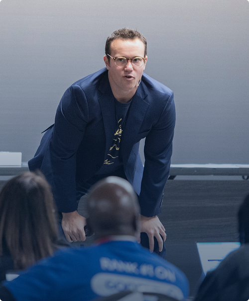 Man in glasses and blue blazer leaning forward while speaking to an audience.