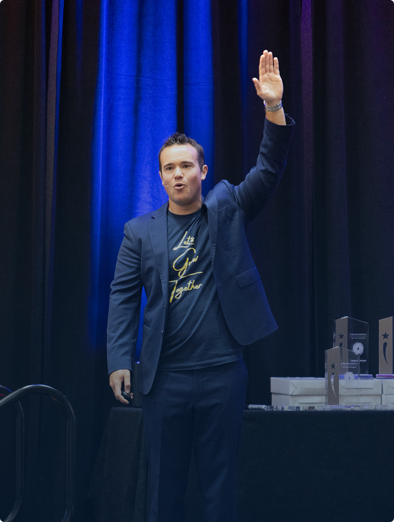 Man in a navy suit and 'Let's Grow Together' shirt speaking on stage with one hand raised.