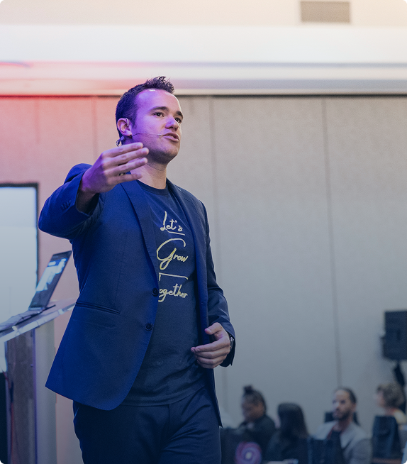 Man in a blazer speaking and gesturing at a podium with an audience in the background.