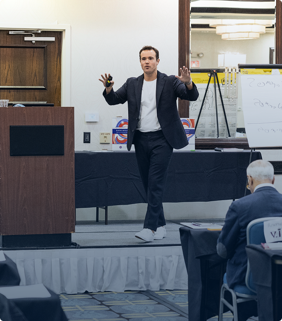 Man in a suit and white sneakers giving a presentation on stage with a whiteboard and audience in the foreground.