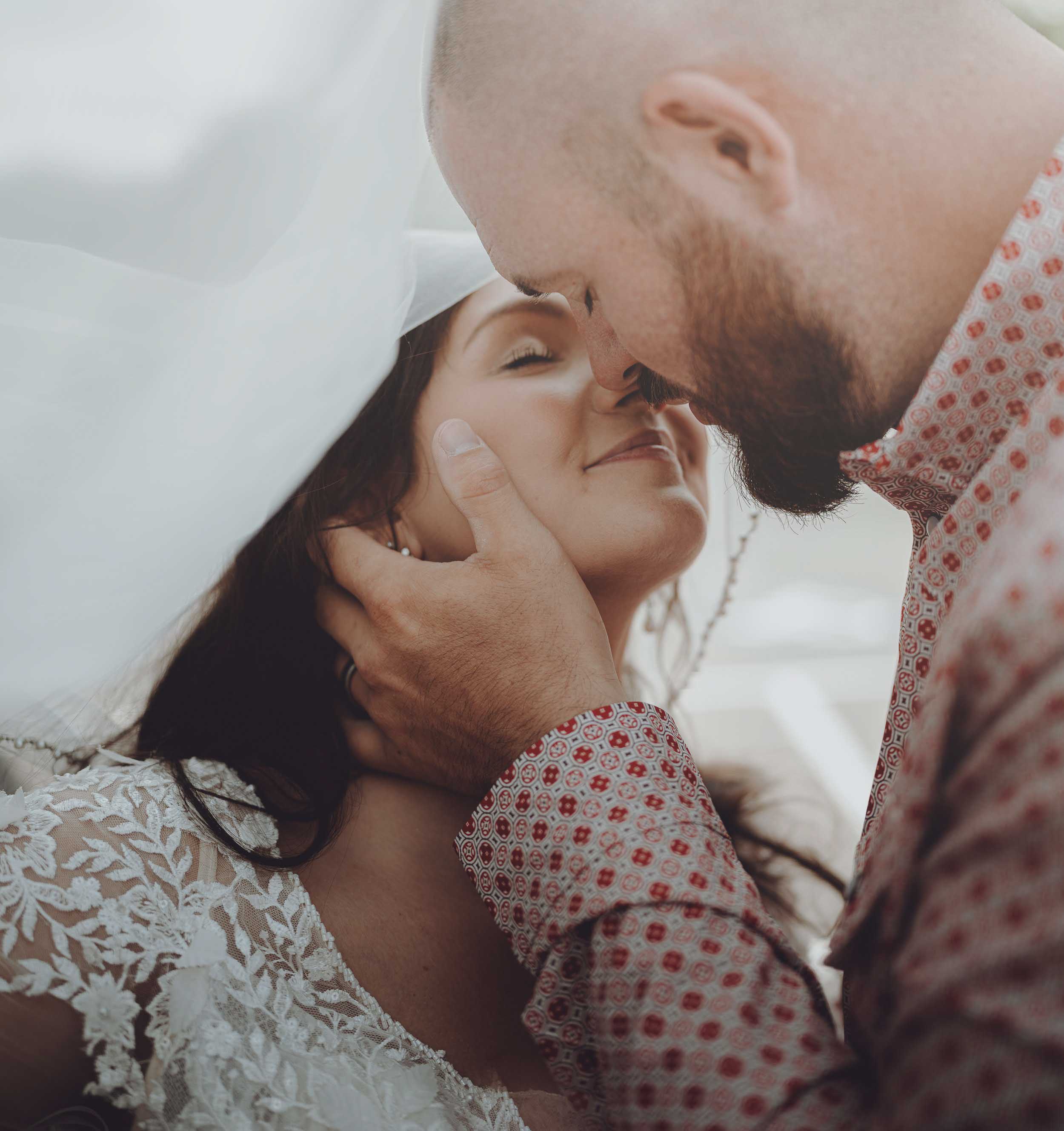 Close-up of a groom tenderly holding the bride’s face as they lean in for a nose touch under her veil.