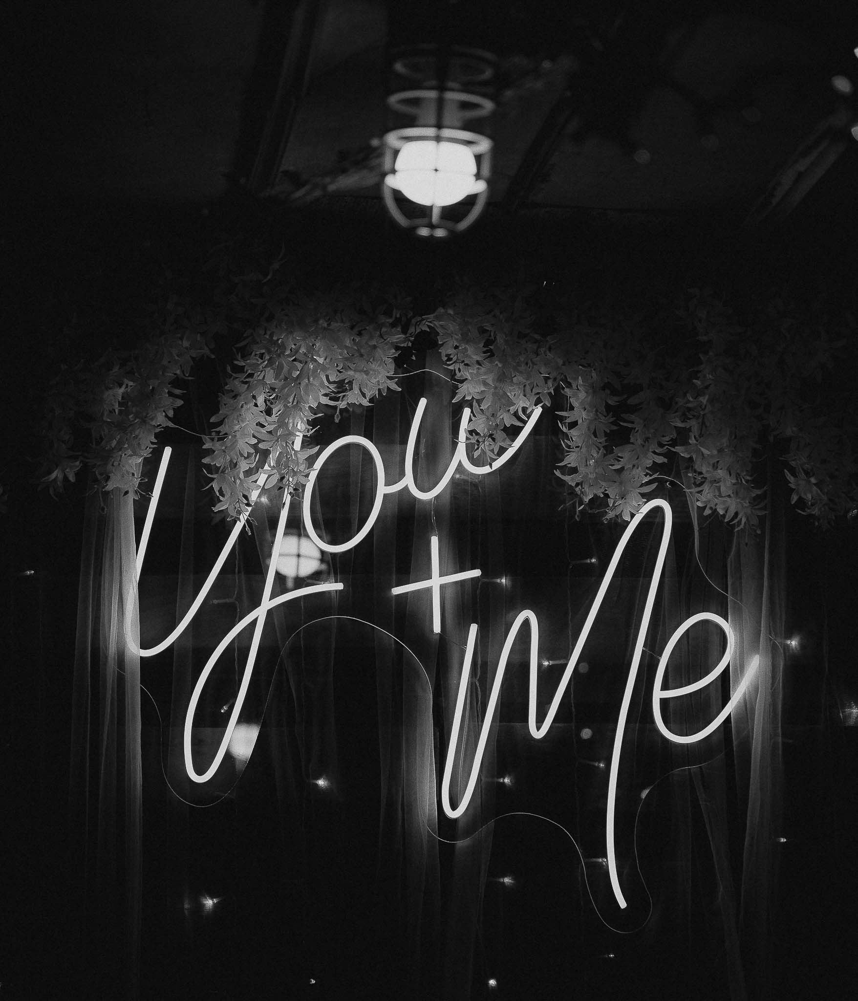 Illuminated neon sign reading 'You + Me' with hanging greenery above in a dark setting.