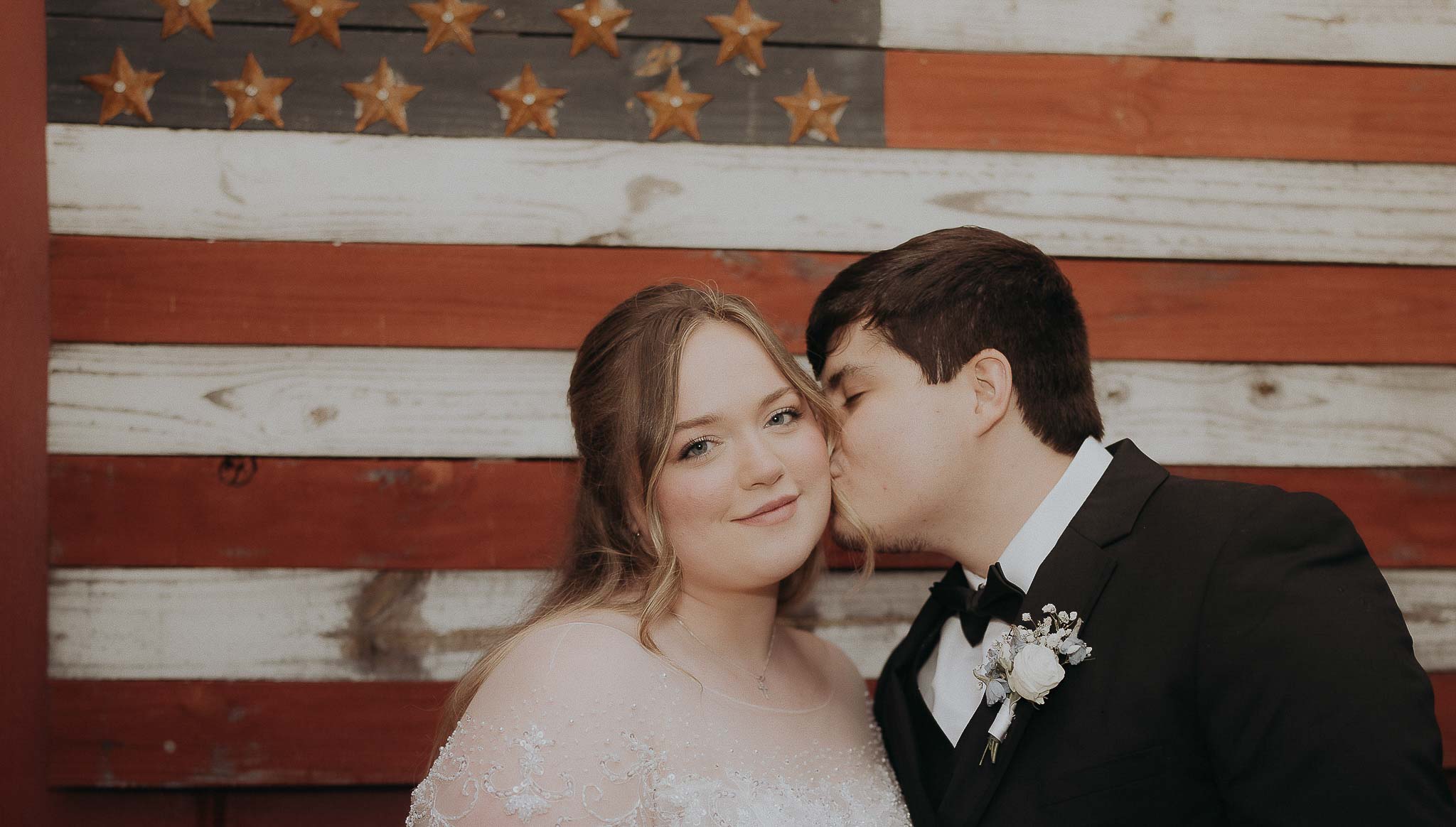 Groom in black tuxedo kissing bride in white lace wedding dress on the cheek in front of a wooden American flag backdrop.