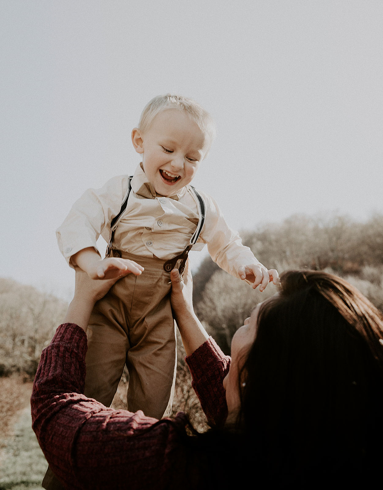 A smiling toddler in a beige shirt and suspenders is lifted in the air by a woman wearing a maroon sweater outdoors.
