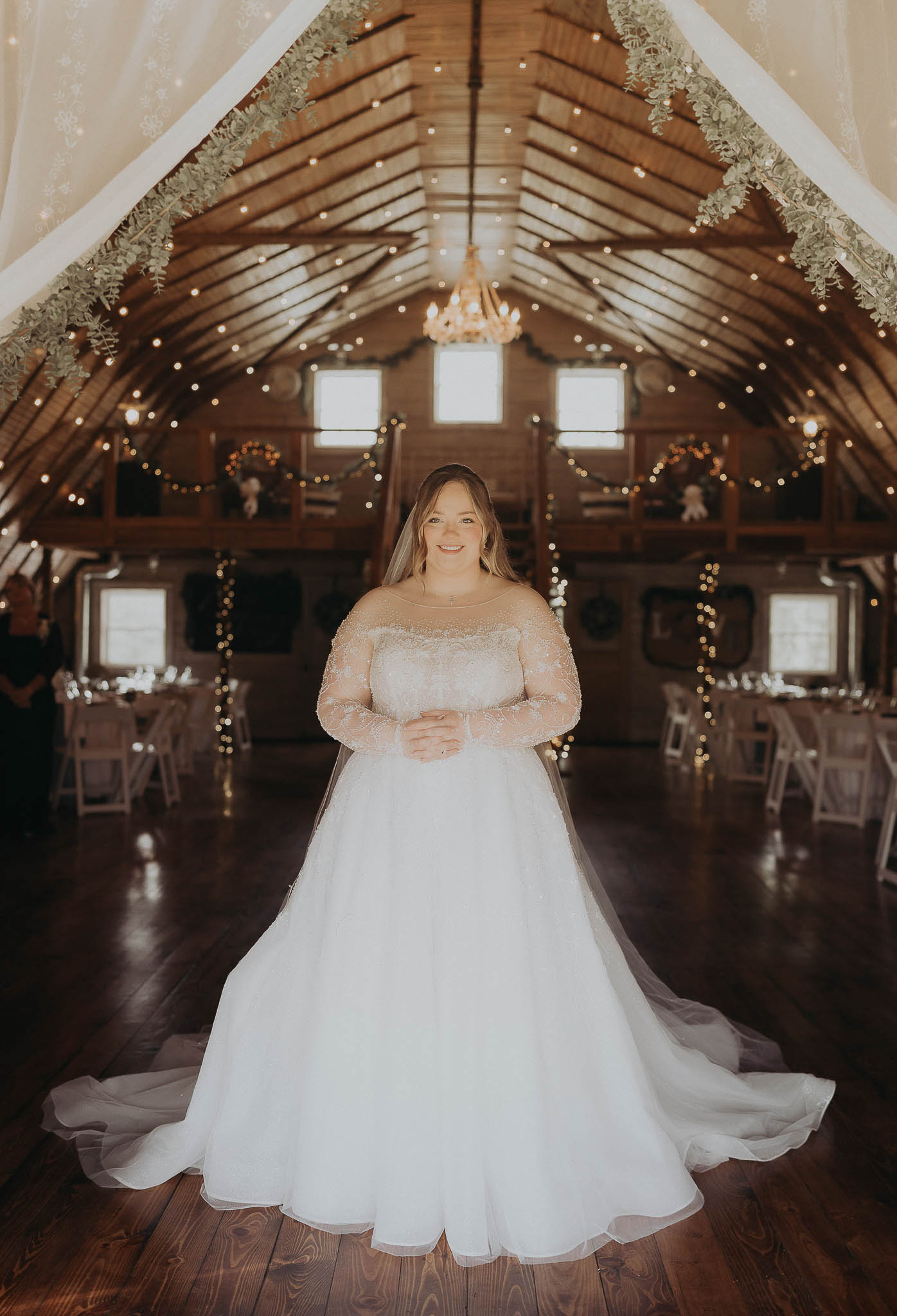 Smiling bride in a white lace wedding dress standing in a warmly lit rustic barn decorated with string lights and greenery.