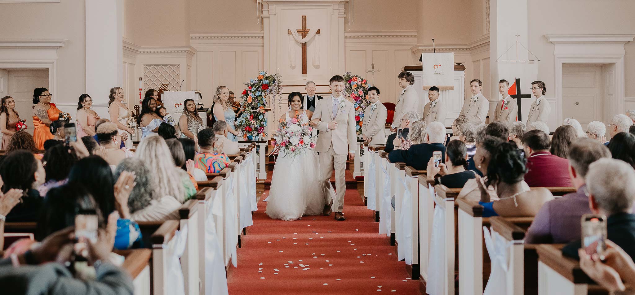 Bride and groom walking down the aisle in a church, surrounded by bridesmaids, groomsmen, and seated guests applauding and taking photos.