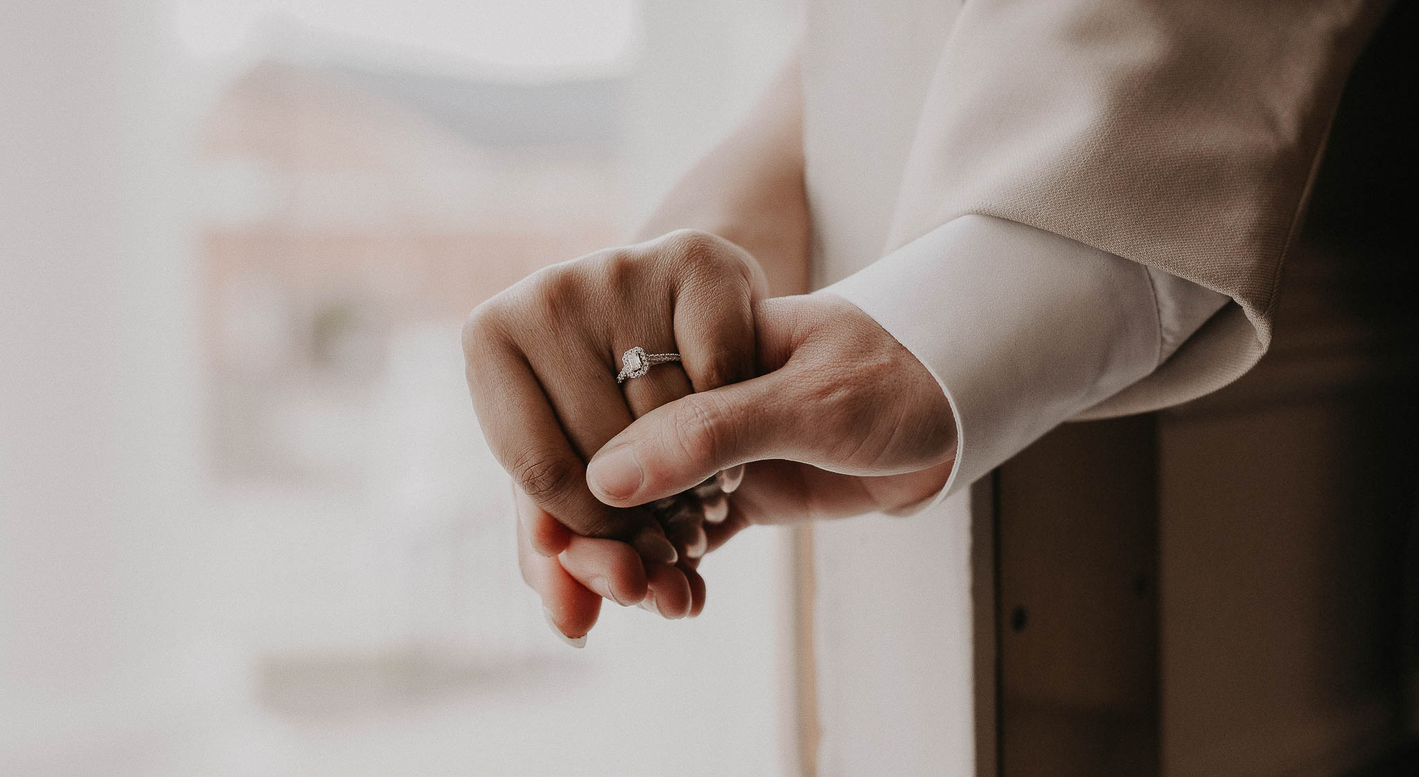 Close-up of two hands gently holding each other, one wearing an engagement ring with a square-cut diamond.