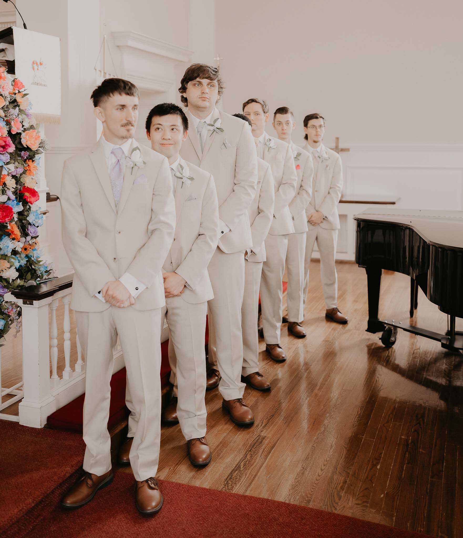 Six groomsmen wearing beige suits and brown shoes standing in a line inside a church near a floral arrangement and a black grand piano.