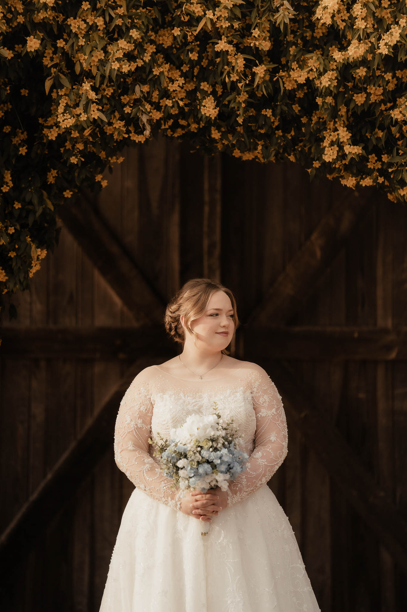 Bride in a white lace wedding dress holding a bouquet of white and blue flowers, standing in front of wooden doors with yellow flowering branches overhead.