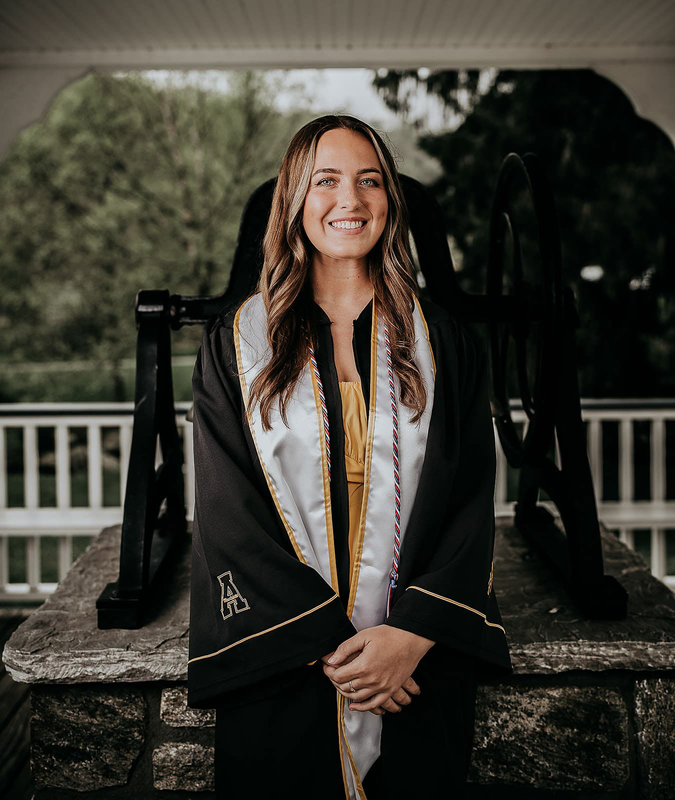 Smiling young woman with long hair wearing a black graduation gown and white stole standing in front of a large bell outdoors.