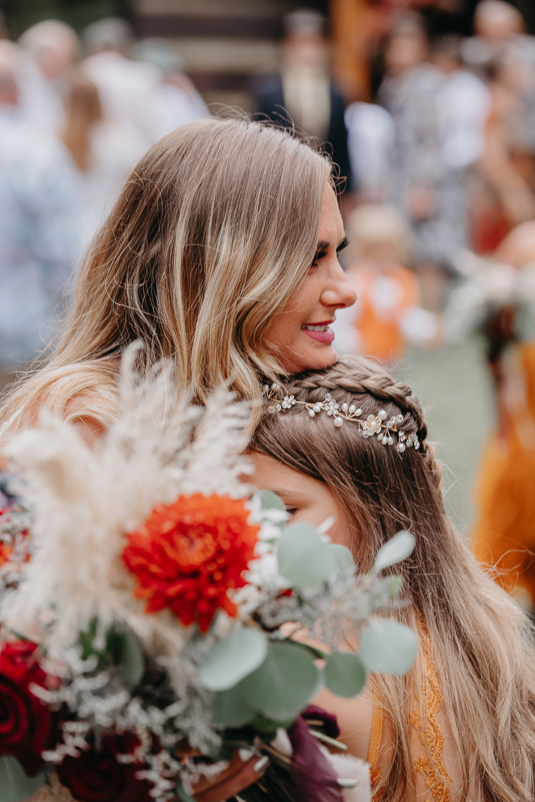 Blonde woman smiling and hugging a girl with braided hair and a floral headpiece, holding a bouquet.