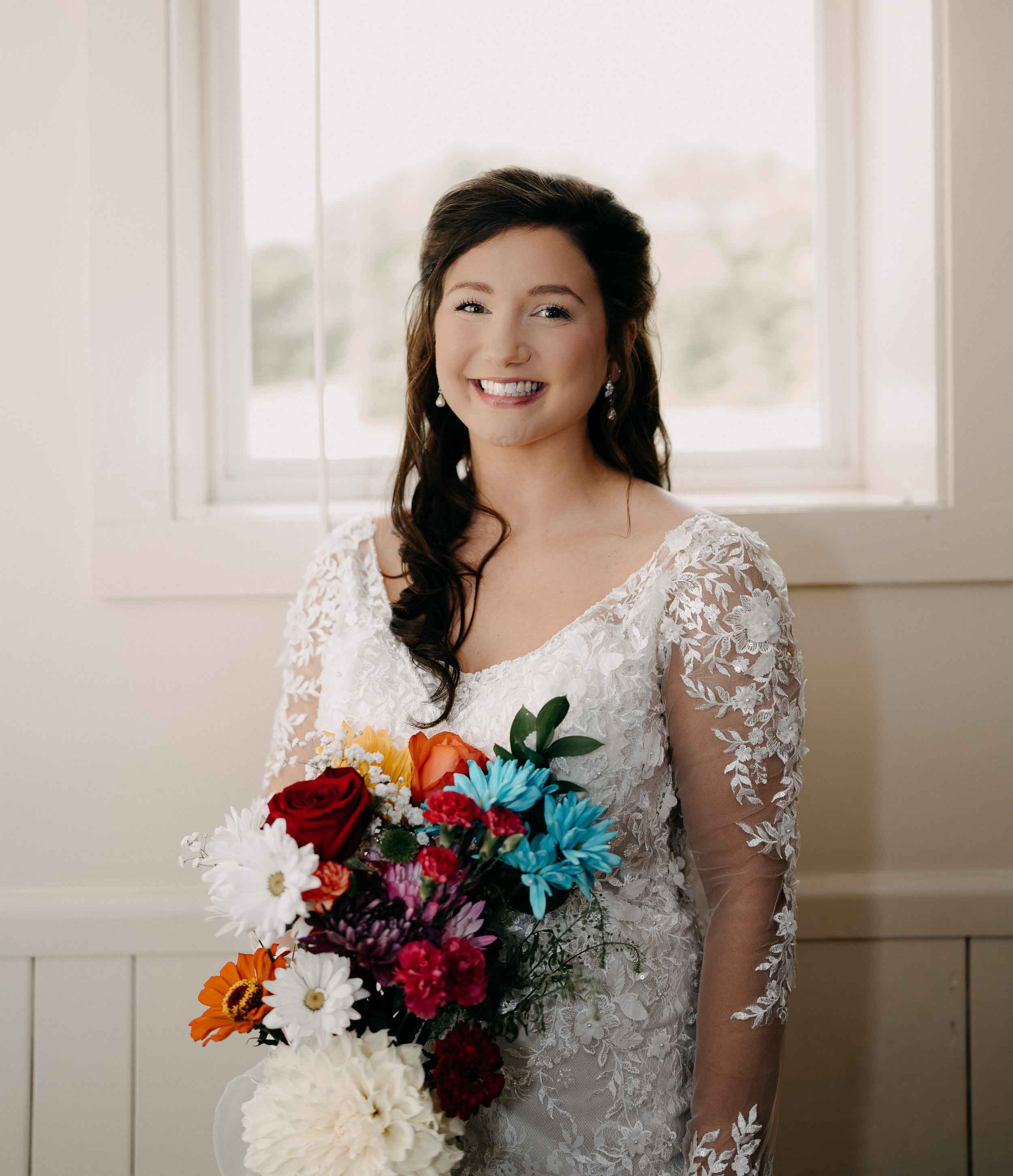 Smiling bride in a white lace wedding dress holding a colorful bouquet of flowers indoors.