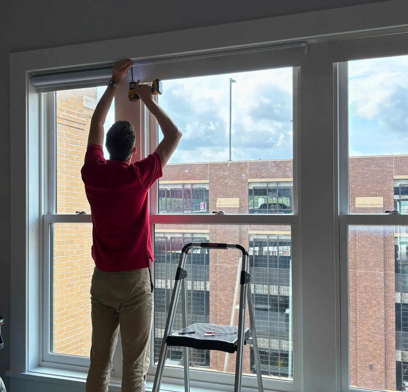 Installer mounting window shades with a drill in front of large city windows