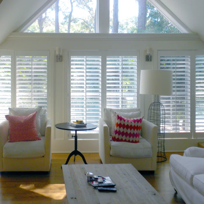 Two tall white plantation shutters installed on windows, with natural light filtering through the louvers into a room with potted plants