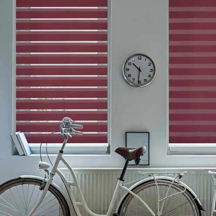 Red zebra shades covering two windows behind a wall-mounted clock and white bicycle