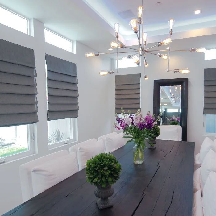 Dark gray Roman shades on tall narrow windows in a formal dining room with white walls and a large chandelier above the table