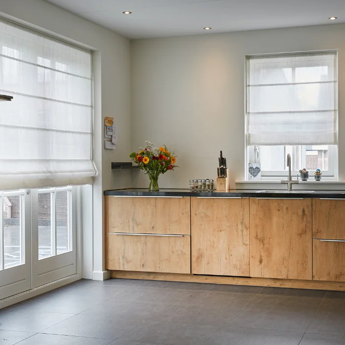 Translucent white Roman shades on two windows in a kitchen with natural wood cabinetry and soft gray walls