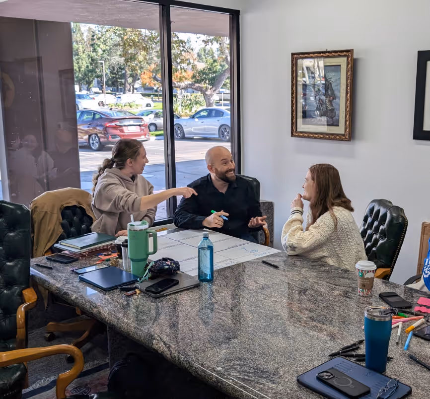 Cole Patterson, founder of heyZensei, laughs and talks with clients around a table with laptops, drinks, and papers in an office.