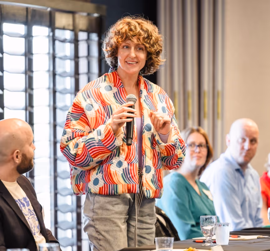 AJ Patterson, founder of heyZensei, is speaking into microphone at a meeting with seated attendees.
