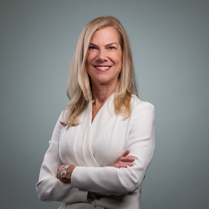 Confident woman with blonde hair in a white blazer smiling with arms crossed against gray background.