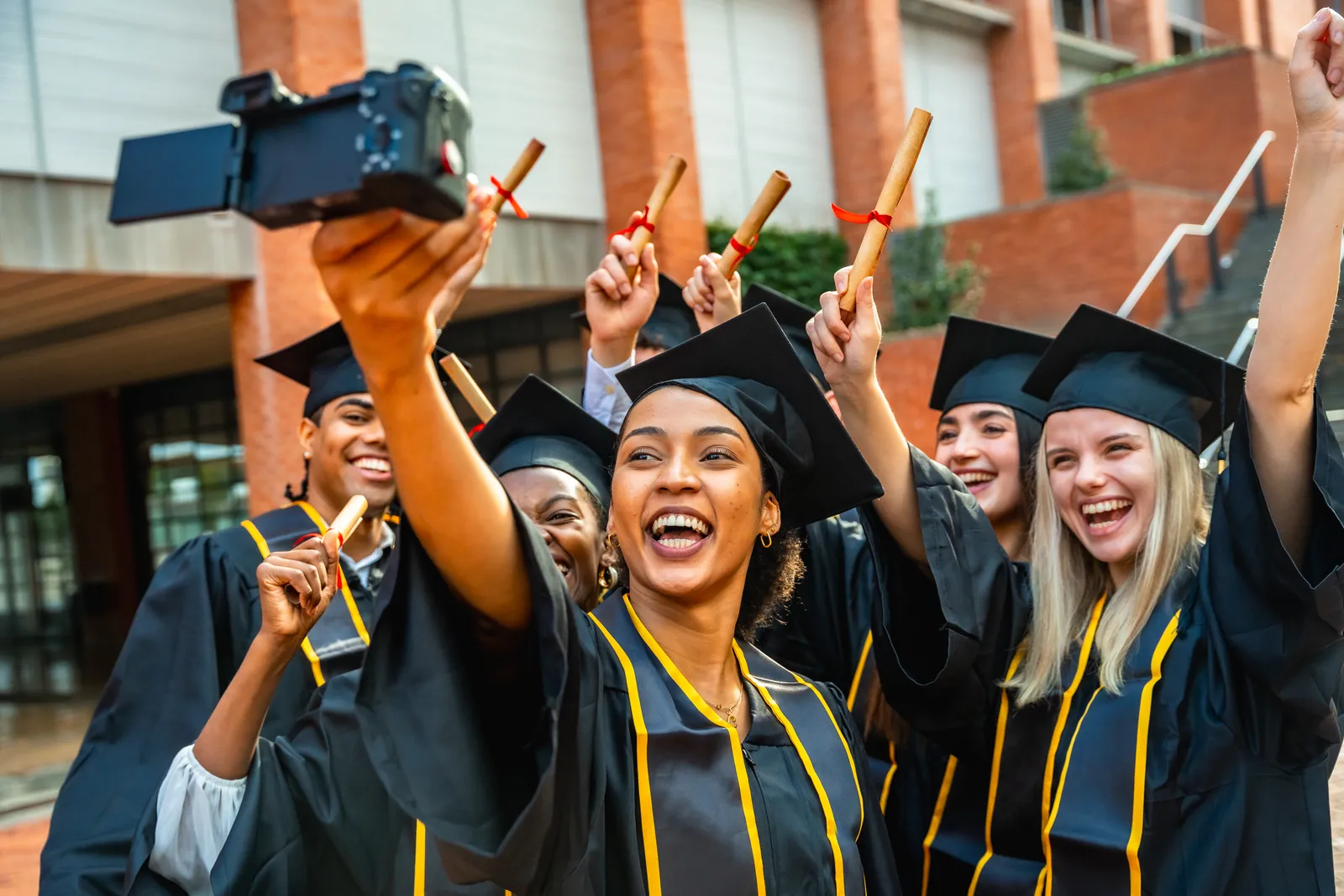 Smiling graduates in caps and gowns holding diplomas and taking a group selfie