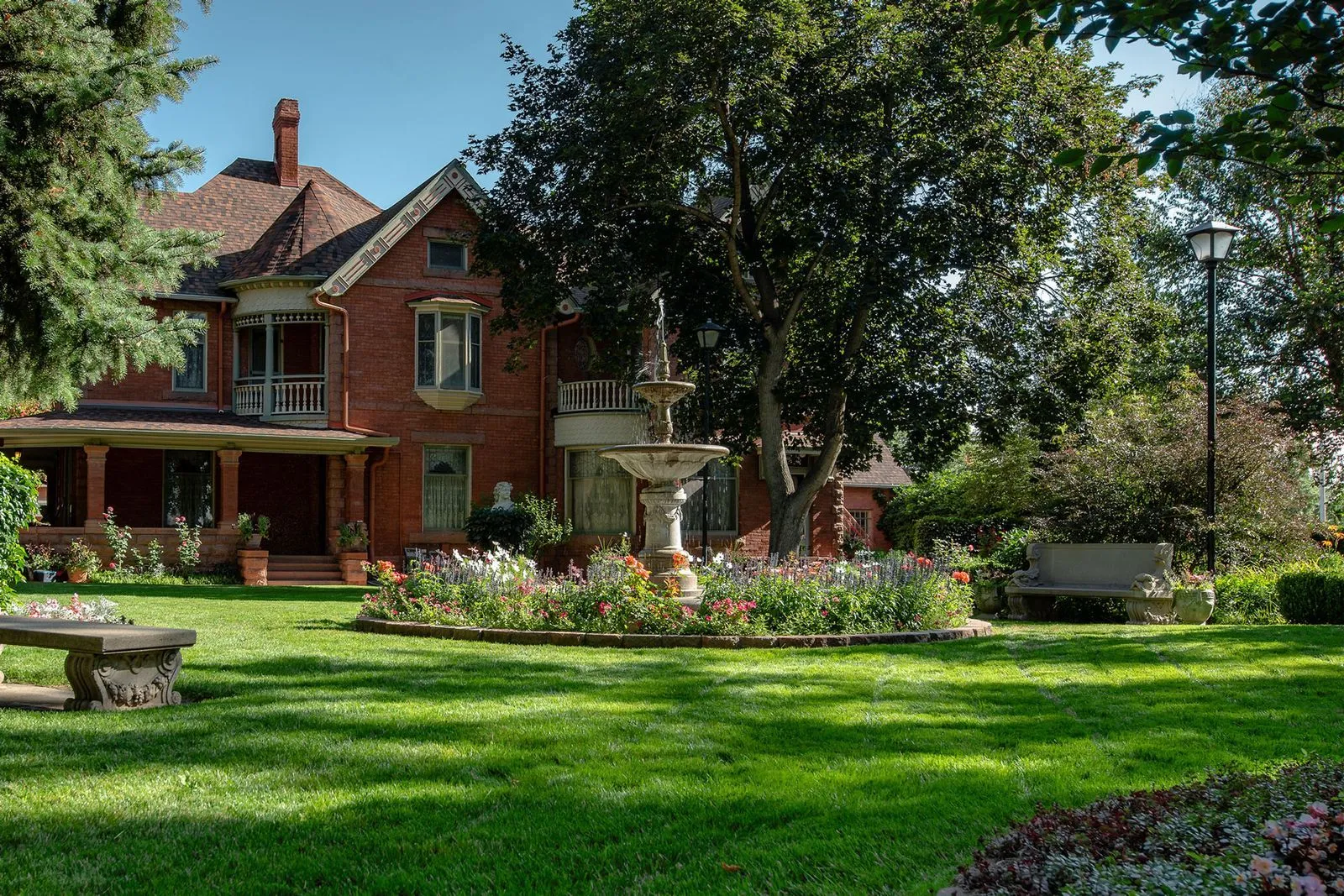 Well-maintained lawn with a brick Victorian house, flowerbeds, a fountain, and a park bench.