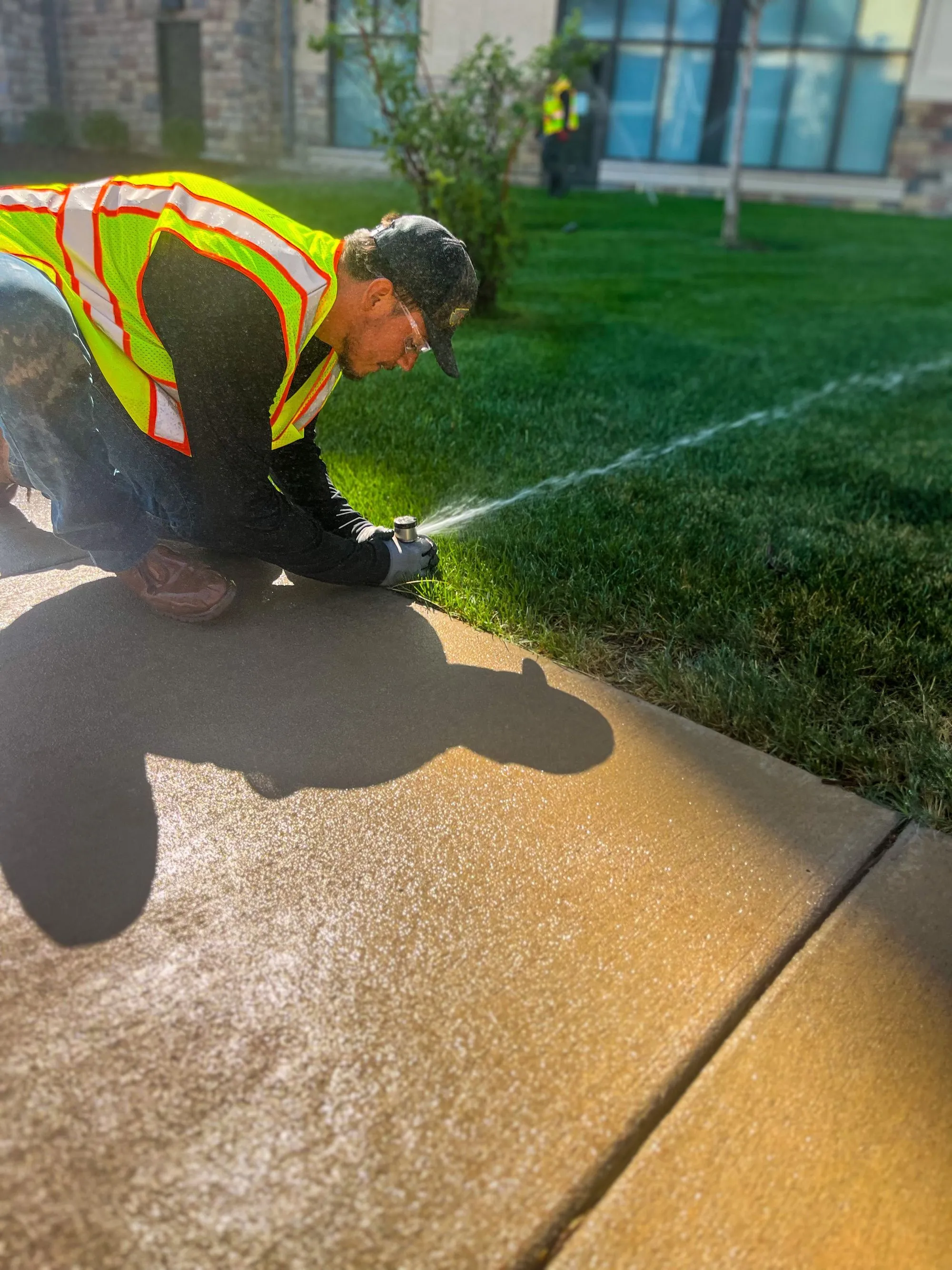 A worker setting up sprinklers for a field of grass beside a narrow concrete pathway. 