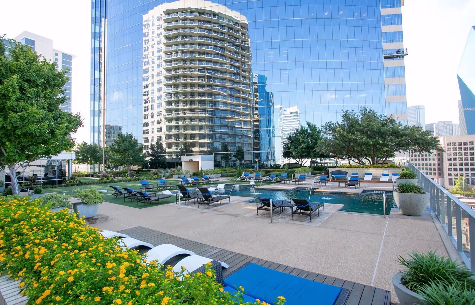 Surrounded by greenery, a rooftop pool sits atop a hotel, bordered by turf, golden blossoms, and neatly placed trees.