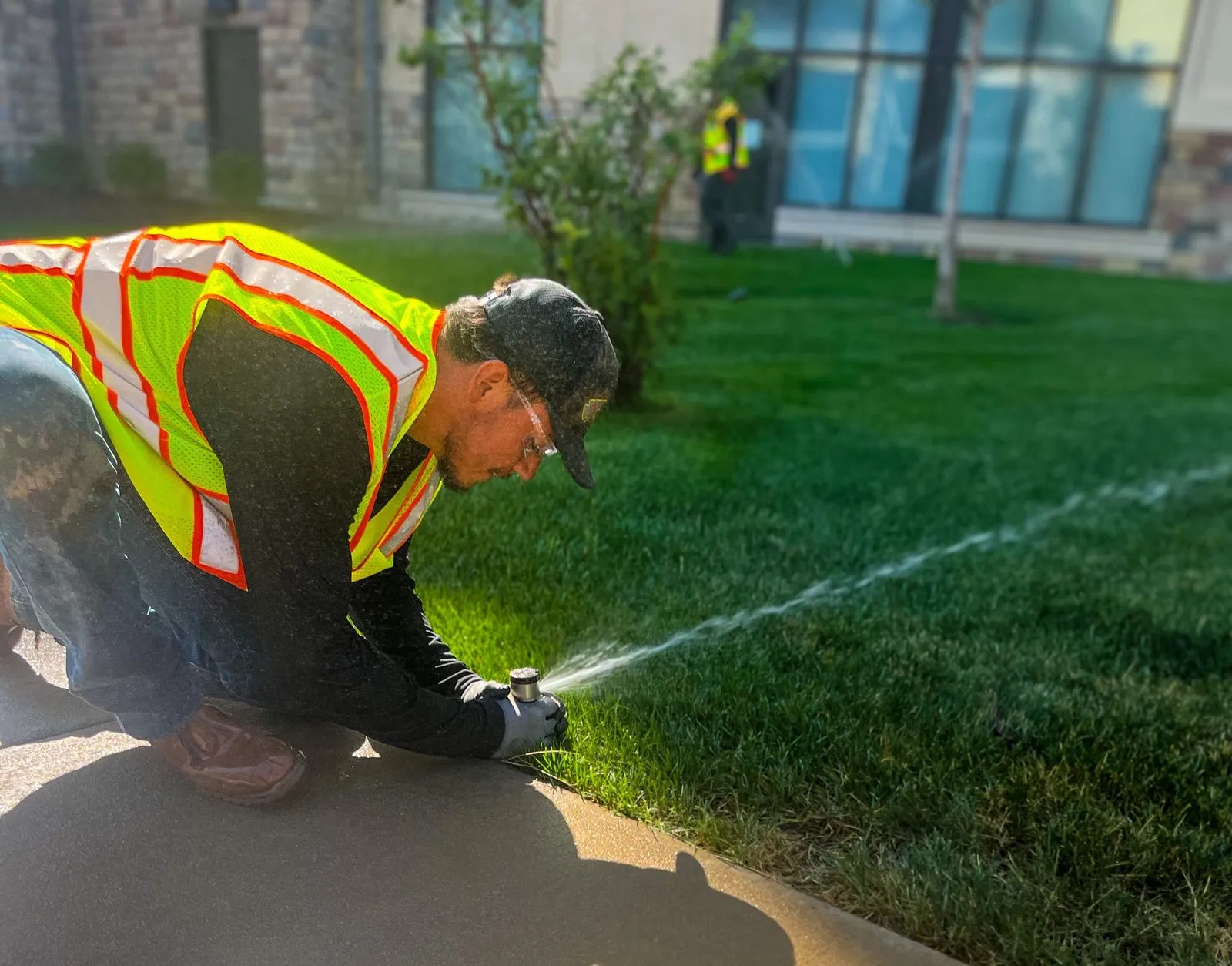 A worker sets up sprinklers across a field of grass, in front of a commercial building. 