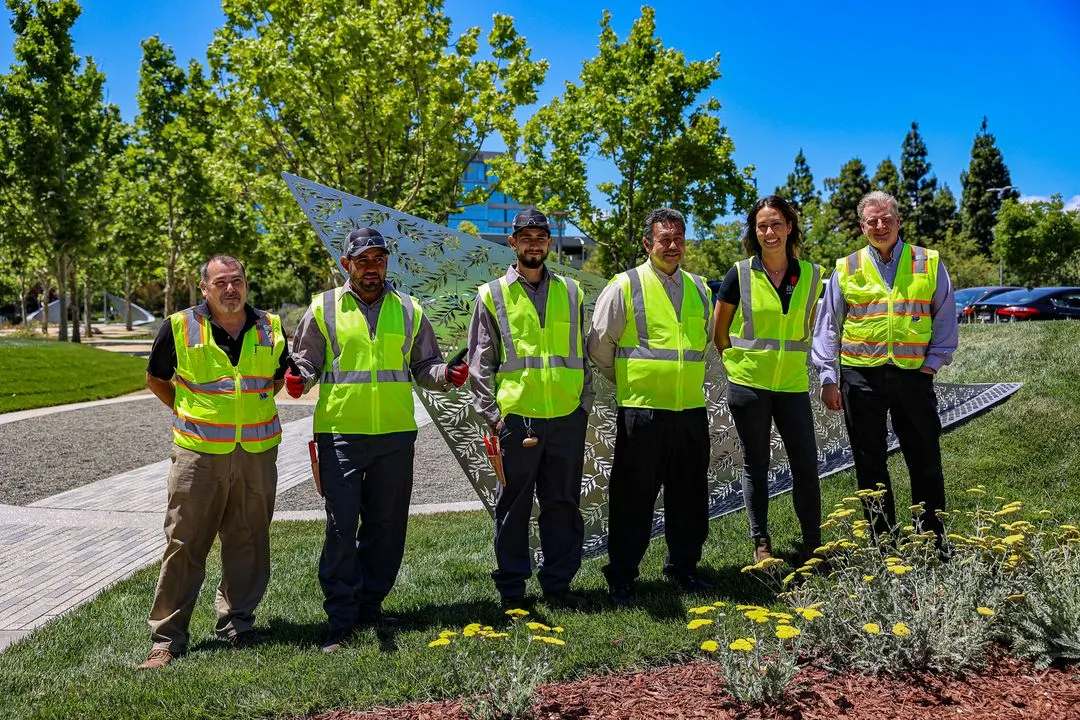 Workers smile in front of a well-maintained landscape of grass and plants aside a commercial building. 