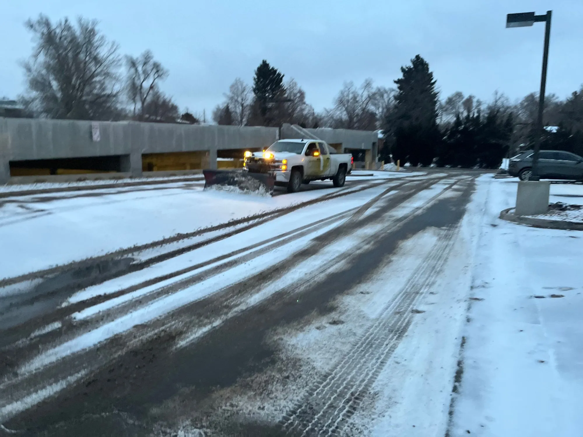 A large truck clears snow from the street running alongside a snow covered parking lot. 