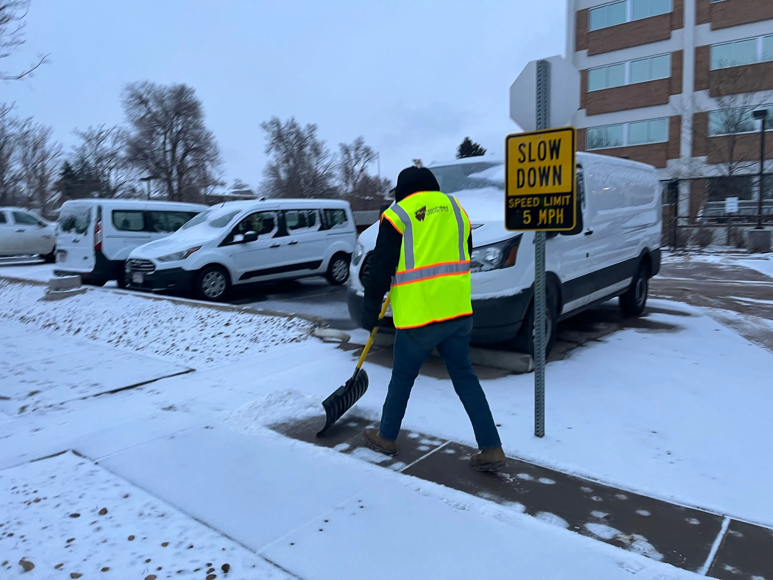 In front of a commercial building, a worker shovels snow from the sidewalk, creating a clear path through the winter morning. 