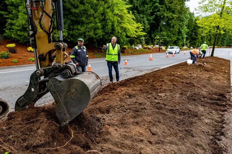 Workers directing an excavator alongside a dirt landscape beside the road.