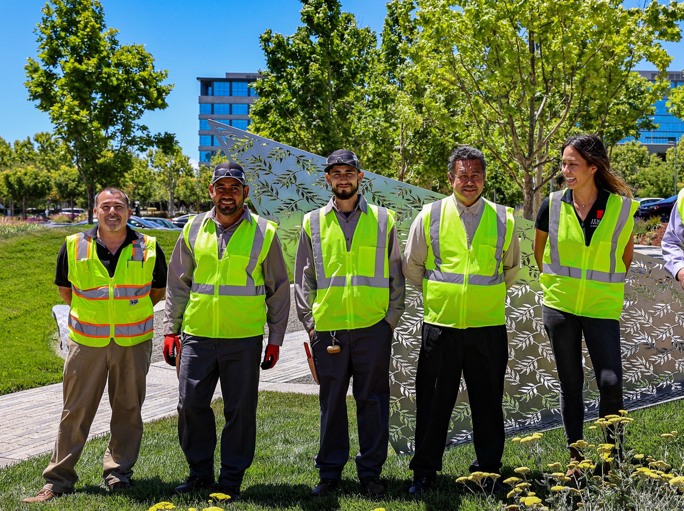 Workers smile in front of a well-maintained landscape of grass and plants aside a commercial building. 