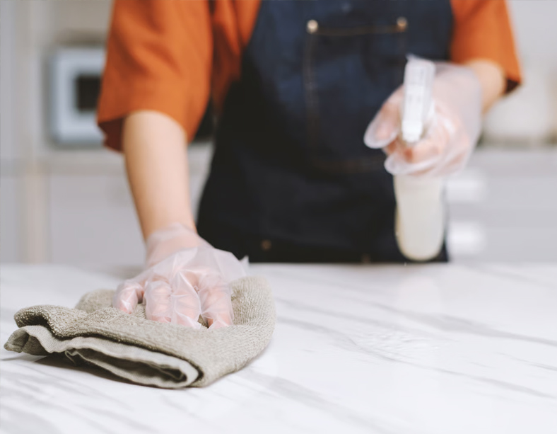 A person safely cleaning and maintaining a natural stone kitchen benchtop using a spray bottle and a microfiber cloth.