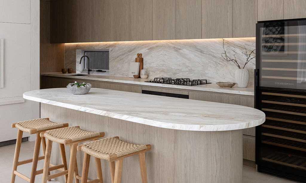 Modern luxury kitchen showcasing a bespoke curved natural stone island benchtop and a matching full-height stone splashback illuminated by LED lighting. The design features ribbed timber cabinetry, woven bar stools, and premium stone surfaces supplied by Marble Hub.