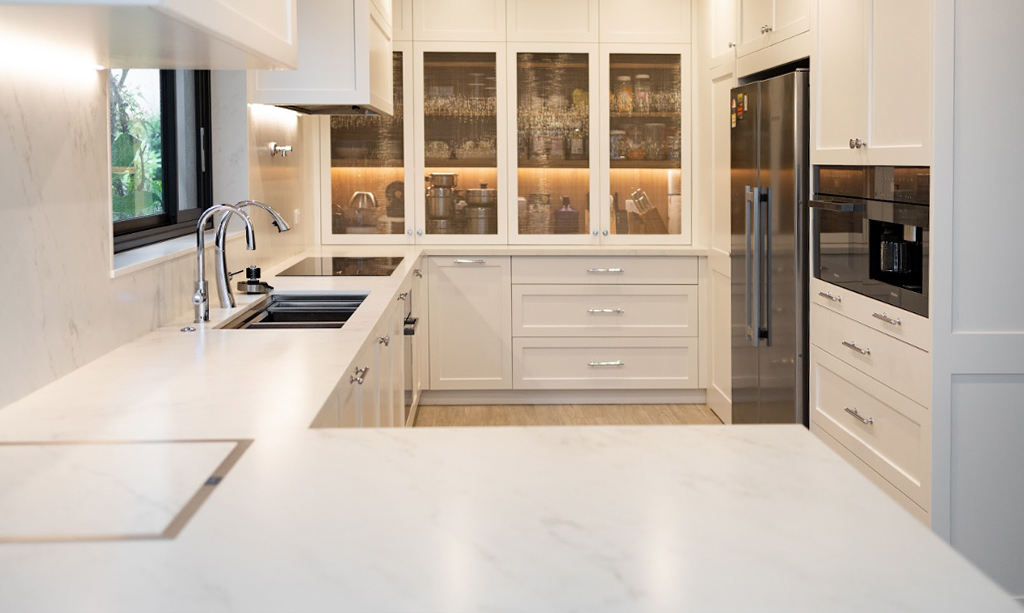 A bright, classic kitchen featuring white cabinetry, backlit glass displays, and a continuous white marble-look engineered stone benchtop and splashback