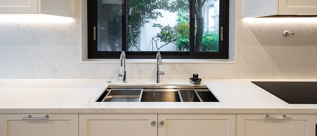Stainless steel undermount double sink and chrome faucets set into a subtle white marble-look engineered stone benchtop below a dark-framed window