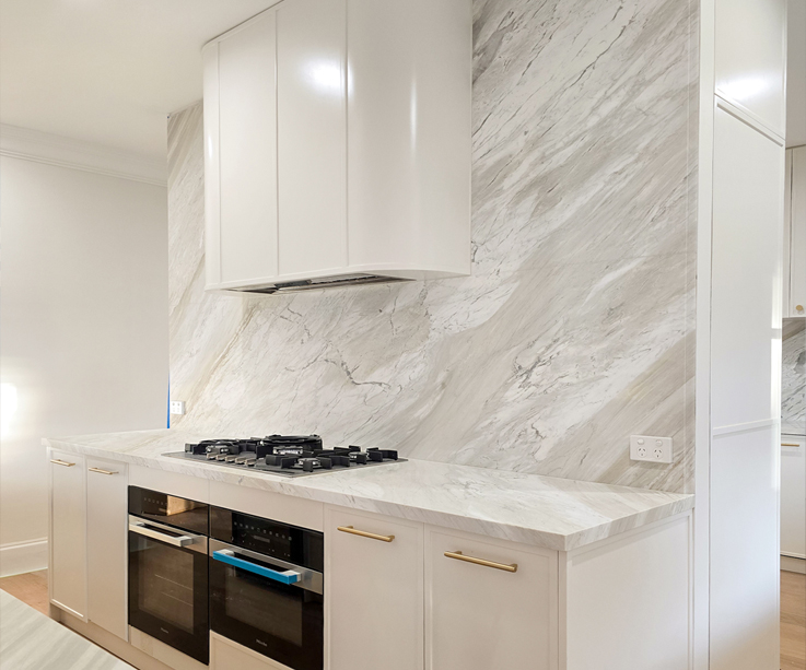 Stone from benchtop to full-height splashback behind the cooktop. The 40mm mitred benchtop edge sits in the foreground.