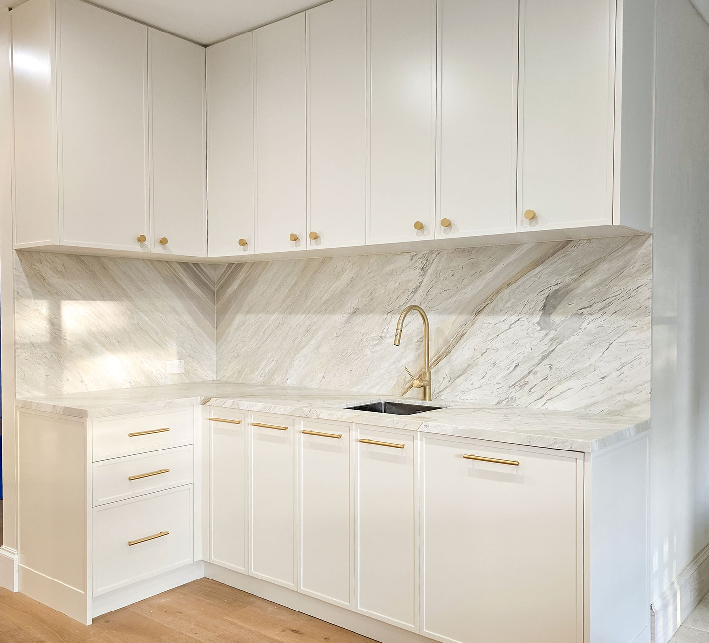 Corner kitchen layout featuring traditional white cabinetry, brass hardware, and a natural stone benchtop with a matching full-height splashback