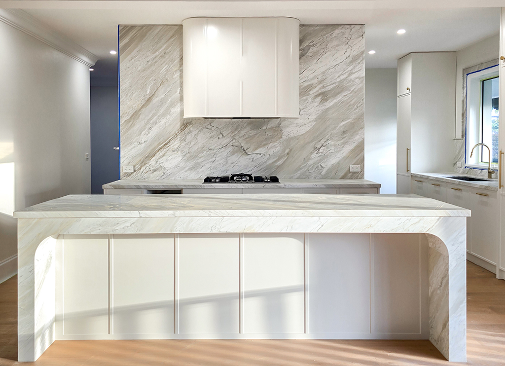 Large natural stone kitchen island with curved waterfall edges, paired with a full-height stone splashback and a custom white rangehood.