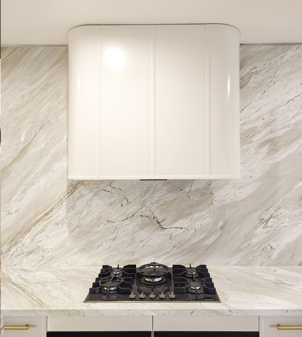 Close-up of a five-burner gas cooktop set into a natural stone benchtop, backed by a continuous stone splashback and a curved white rangehood