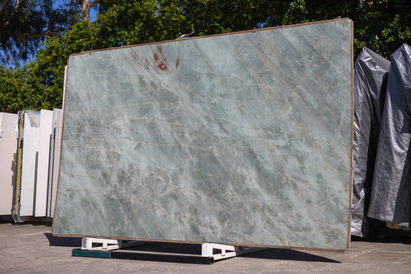 A full-size unpolished blue-green quartzite stone slab with intricate grey and red-brown veins, supported by a metal frame stand. Displayed in an outdoor stone yard with dark green trees and a clear blue sky in the background
